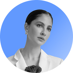 Black and white portrait of a woman with slicked-back hair wearing earrings and a white blazer, holding a flower, on a blue circular background.
